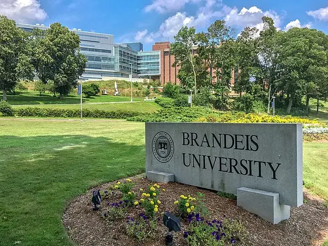 A stone sign reading Brandeis University stands in front of a landscaped lawn with flowers, trees, and modern academic buildings in the background under a partly cloudy sky.