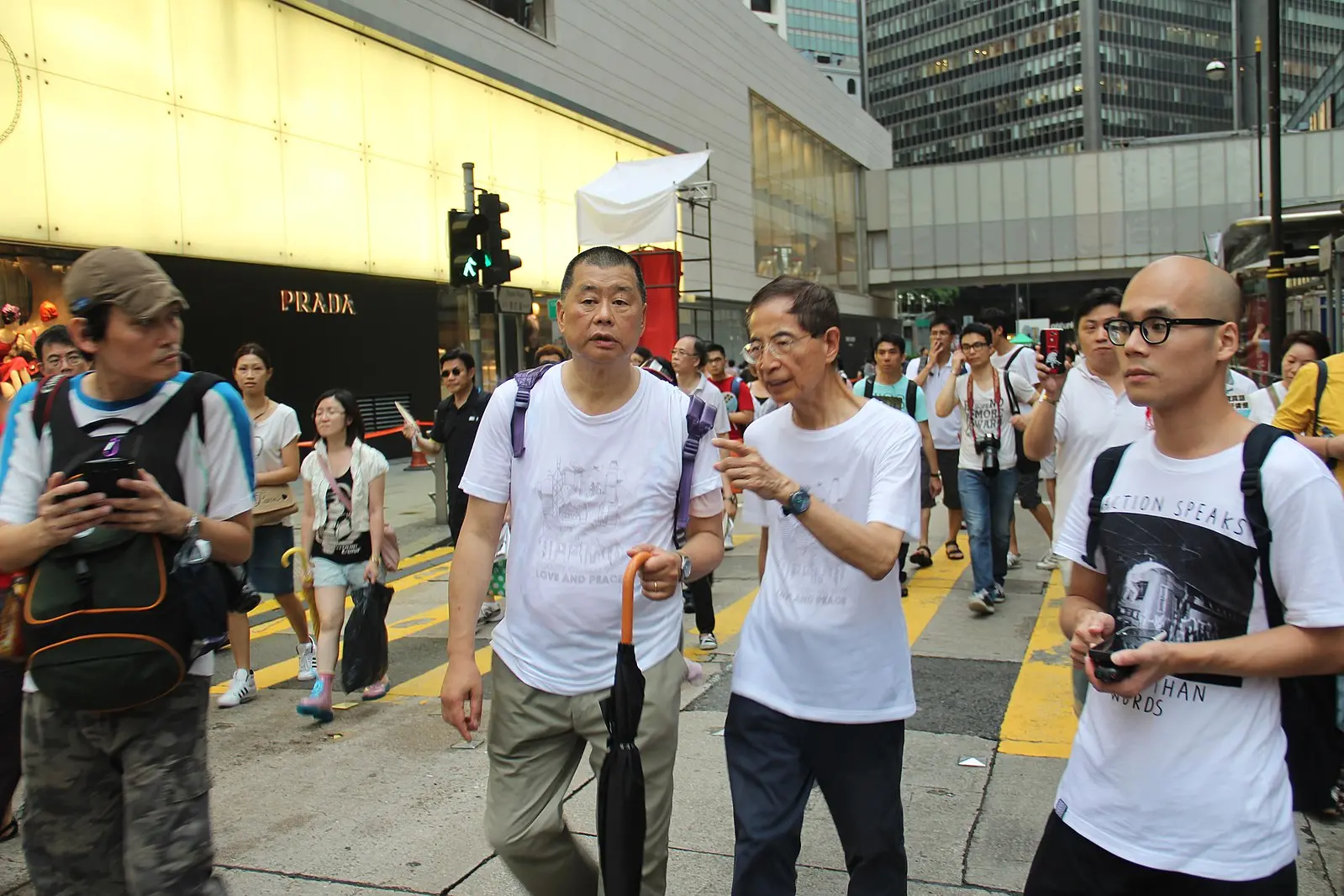 A crowd of people cross a busy city street, with several men in white T-shirts walking in the foreground. Tall buildings and a Prada storefront are visible in the background.