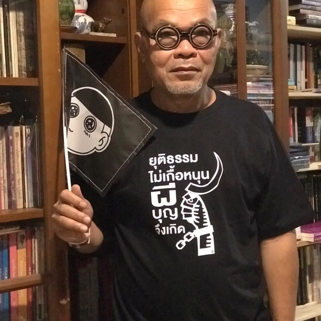 A man with round glasses stands in front of bookshelves, holding a flag with a cartoon face. He wears a black T-shirt with white Thai text and a graphic of a key and hand.