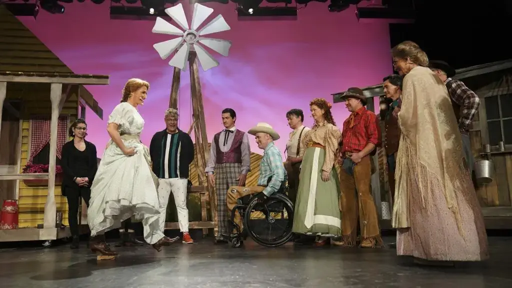 A group of actors in colorful costumes performs a scene on stage, with a windmill and farmhouse set in the background. One woman stands center, facing a man in a wheelchair, surrounded by others watching intently.