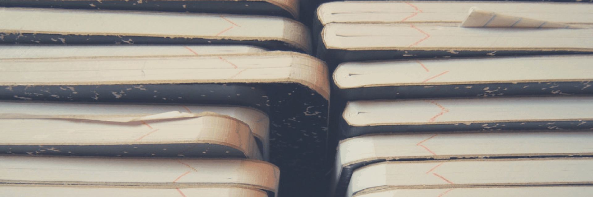 Close-up photo of stacked composition notebooks with black spines and worn, white covers. Some pages are slightly bent or sticking out, showing signs of frequent use.