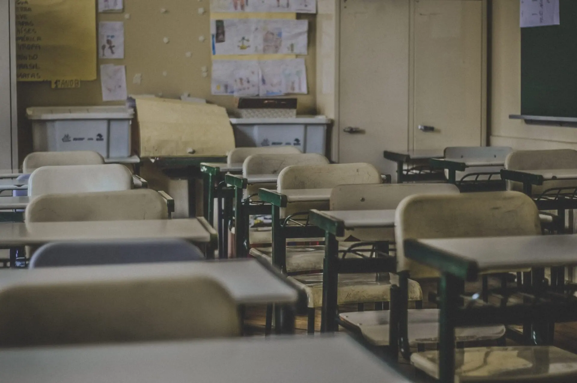 Empty classroom with rows of desks and chairs, papers on the walls, cabinets, and a chalkboard in the background. The room appears dimly lit and quiet, with no students or teachers present.