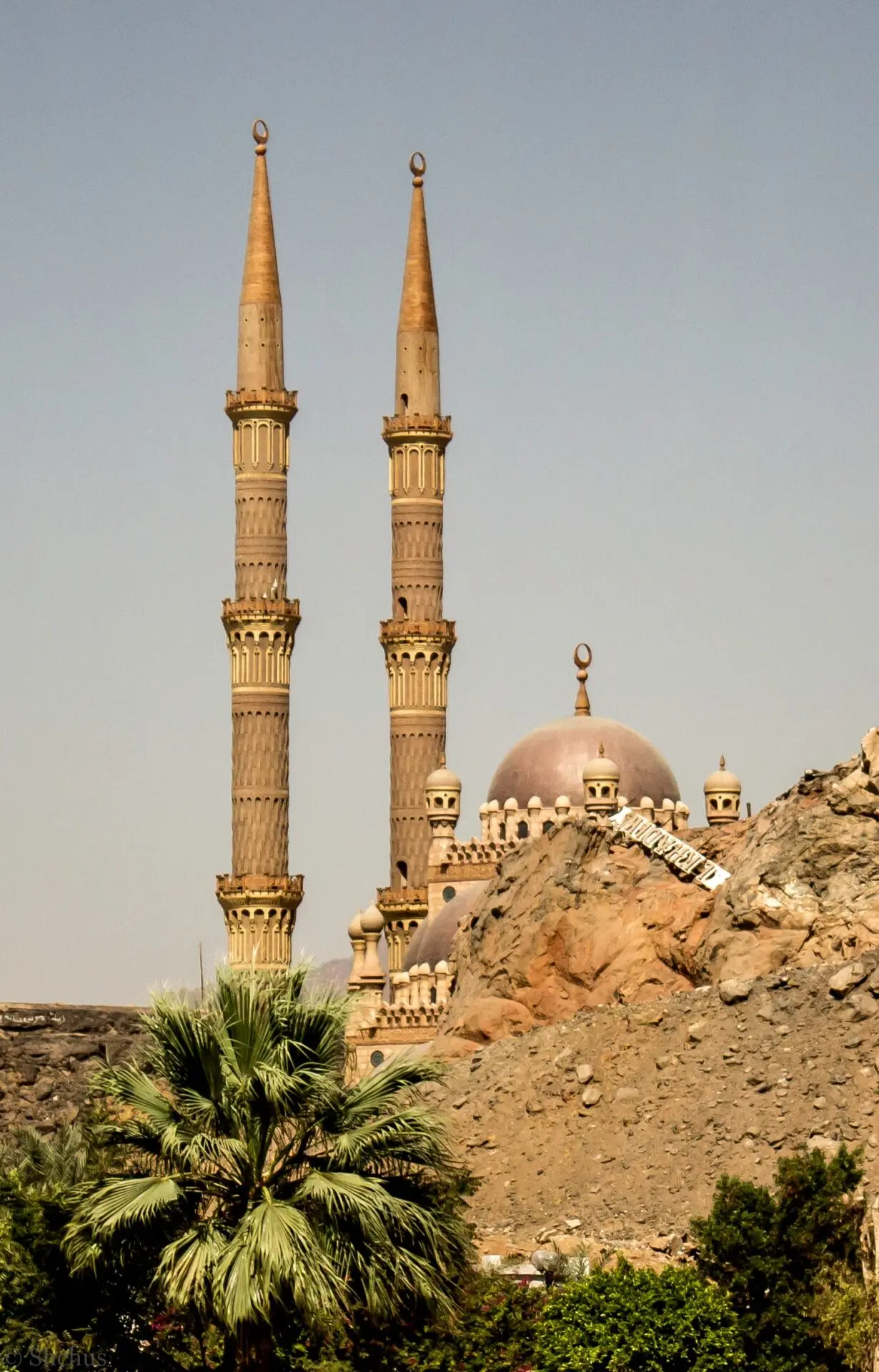 A mosque with two tall, ornate minarets and multiple domes stands behind a rocky hill, surrounded by greenery and palm trees under a clear sky.