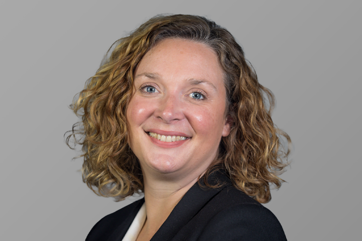 A woman with curly, shoulder-length light brown hair, wearing a black blazer and white top, smiles at the camera against a plain grey background.