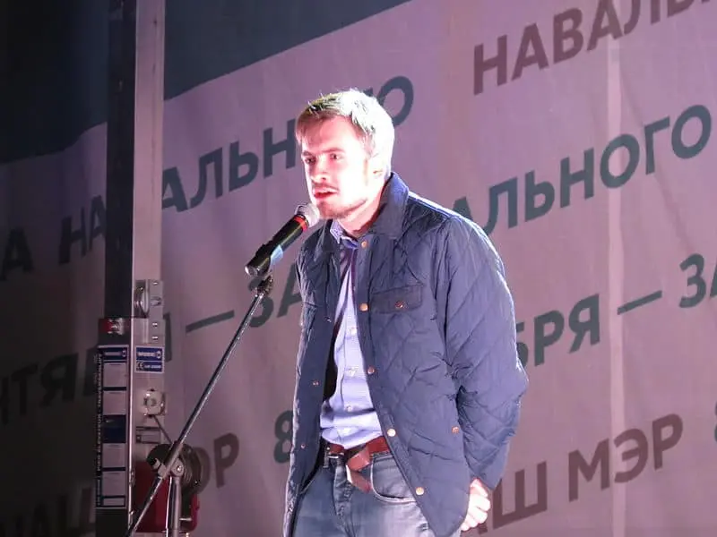 A man in a blue jacket stands and speaks into a microphone on stage. Behind him is a banner with Russian text and the name Navalny prominently visible.