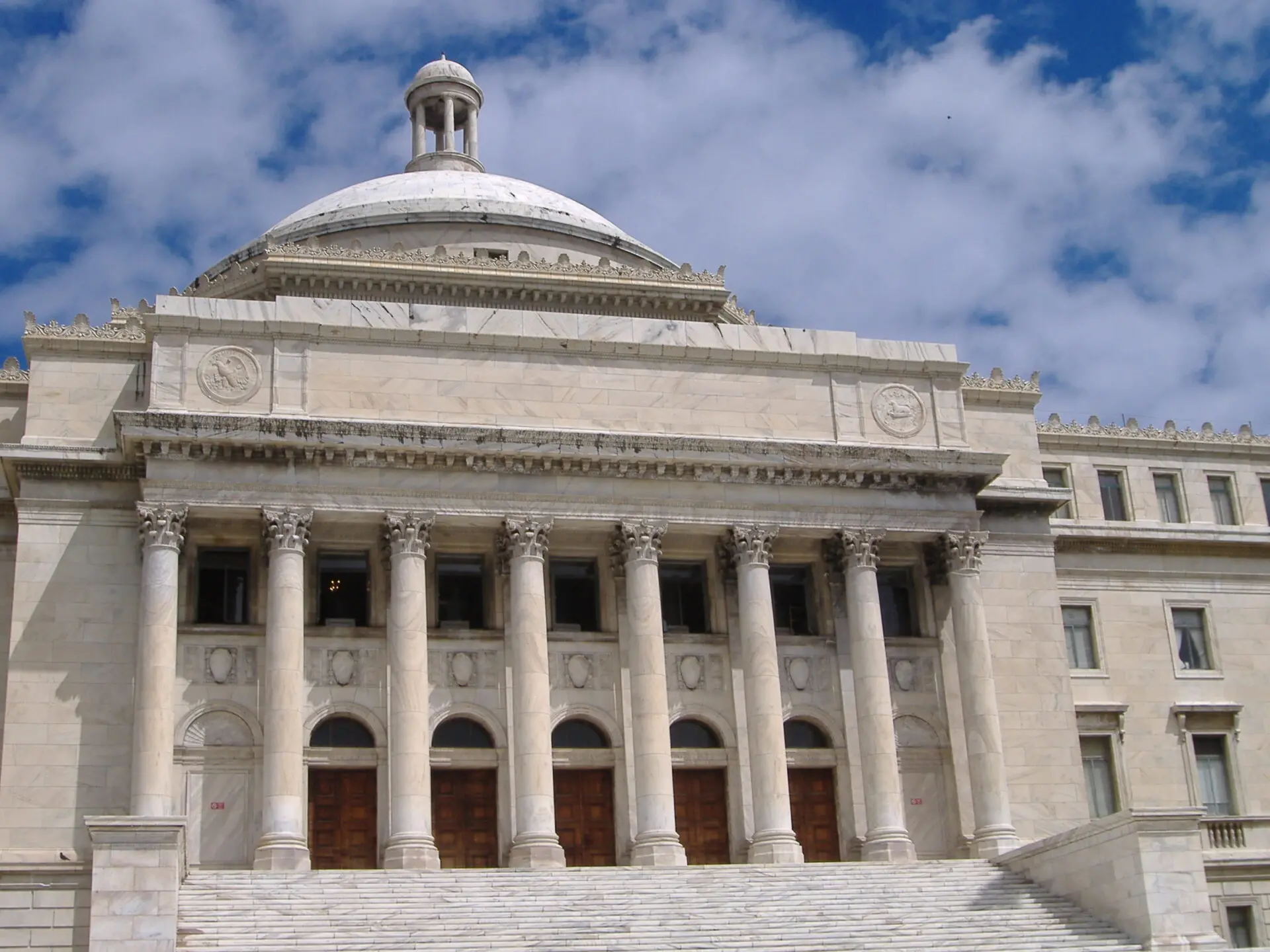 A neoclassical government building with a large dome, tall columns, and wide steps leading to multiple wooden doors, set against a partly cloudy blue sky.