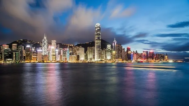 A vibrant city skyline with tall, brightly lit skyscrapers reflecting on a body of water at dusk, under a dramatic, cloud-filled sky.
