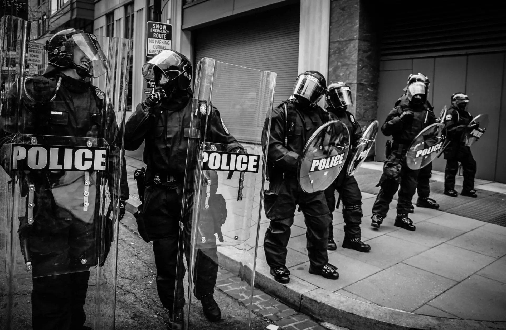 Five police officers in riot gear and helmets stand in a row on a city sidewalk, holding large transparent shields labeled POLICE. The scene appears tense and serious.