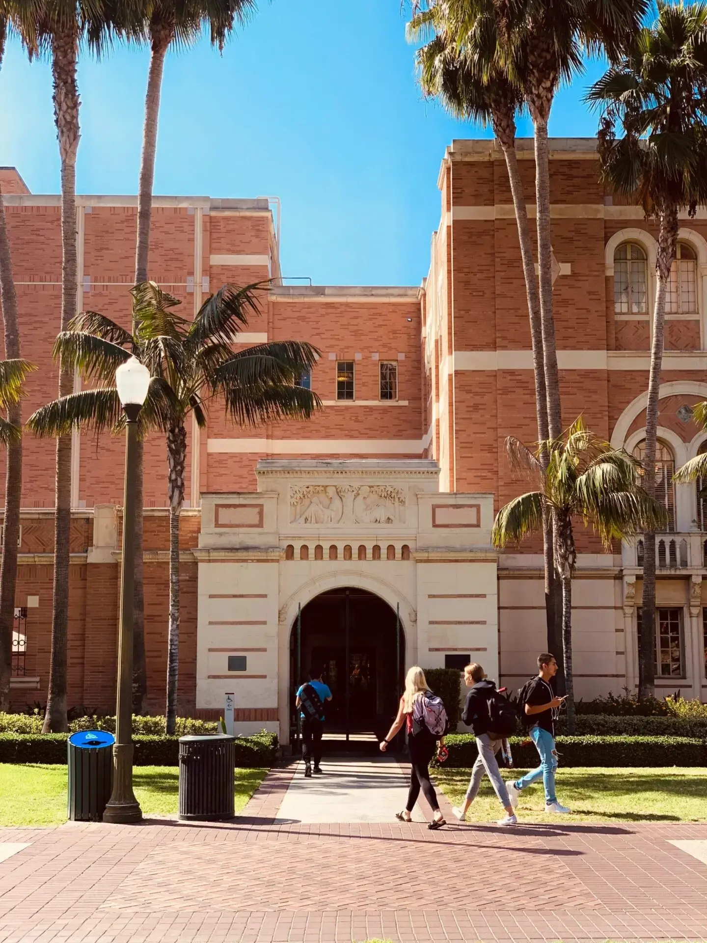 Students walk toward the entrance of a large brick university building lined with tall palm trees on a sunny day. A lamp post, trash bins, and green lawn are visible in front of the building.