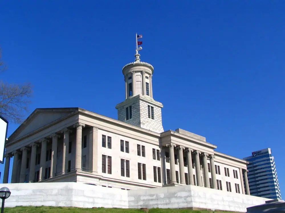 A neoclassical government building with tall columns and a central tower stands against a clear blue sky, with a modern office building visible in the background.