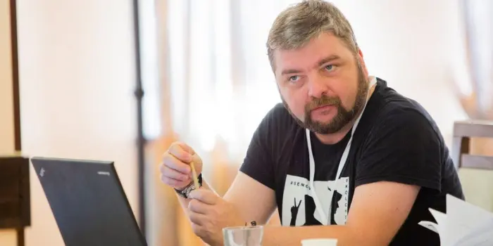 A man with short hair and a beard sits at a table with a laptop, holding an object in his hand, and looking to the side with a thoughtful expression. The setting appears to be indoors, possibly at a meeting or conference.