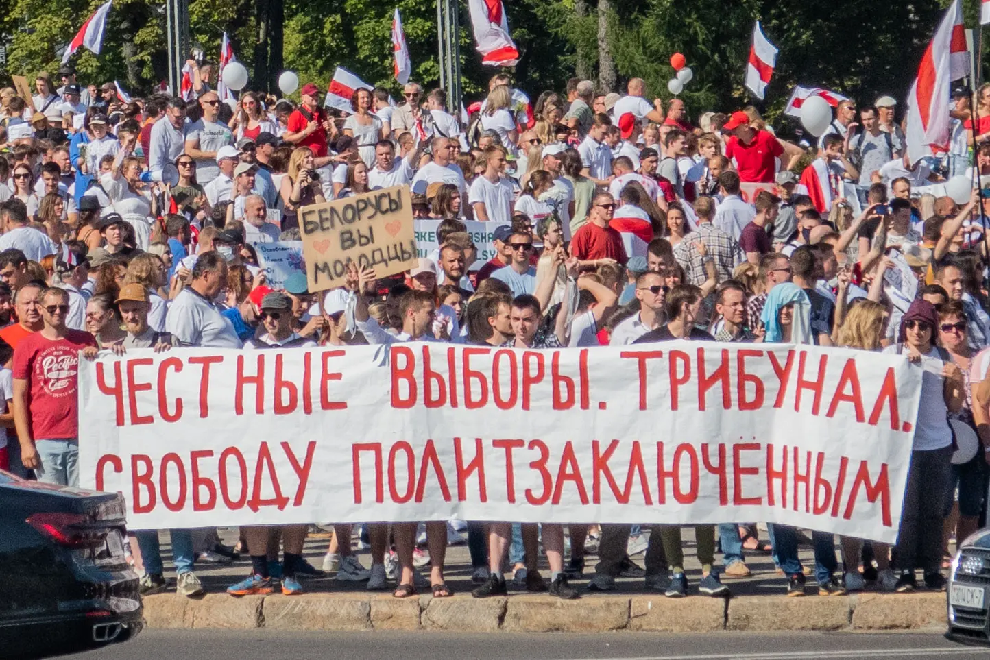 A large crowd holds a banner in Russian calling for fair elections, a tribunal, and freedom for political prisoners. People wave flags and hold various signs during a protest on a sunny day.