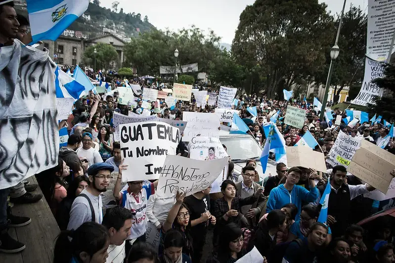 A large crowd protests outdoors, many holding signs and Guatemalan flags. Some signs are handwritten with messages; one reads GUATEMALA Tu Nombre and another No Tenemos Presidente. Trees and buildings are in the background.