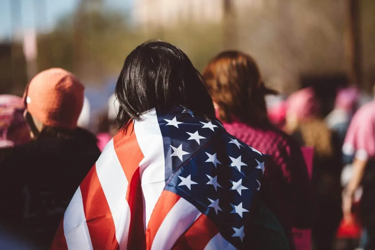 A person draped in an American flag stands among a crowd outdoors, where several others in pink hats and jackets suggest a protest, possibly against a tax.