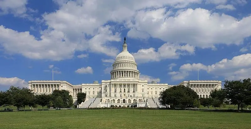 The United States Capitol building in Washington, D.C., with its white dome and columns, stands under a partly cloudy blue sky—an enduring symbol of democracy and recent coronavirus relief efforts—viewed from a grassy lawn with trees on both sides.