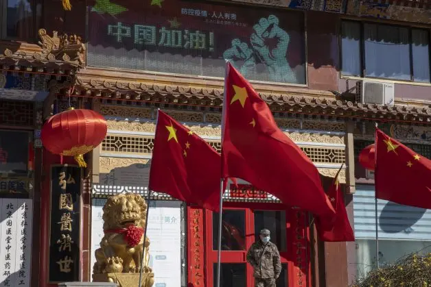 Three Chinese flags and red lanterns adorn a building with traditional decorations and a stone lion statue, while a person in a face mask stands outside—reflecting the ongoing impact of coronavirus. Chinese characters and a digital display are visible behind.