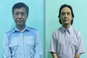 Two men are standing against a plain light blue background, each facing the camera. The man on the left wears a light blue shirt with darker blue shoulders, and the man on the right wears a light purple short-sleeved shirt.