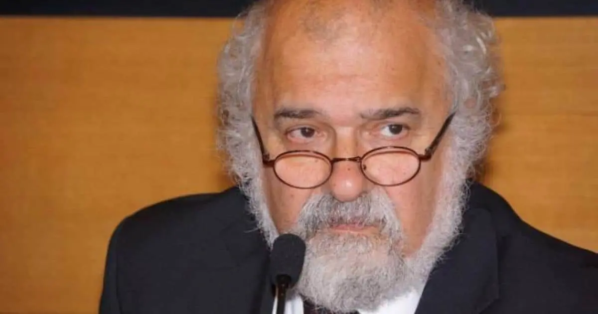 An older man with gray curly hair, a beard, and glasses is wearing a suit and sitting in front of a microphone, looking serious. The background is a solid brownish color.