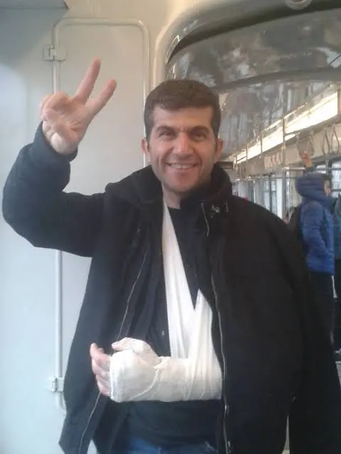 A man with a bandaged and sling-supported arm smiles and flashes a peace sign while standing inside a public transportation vehicle.