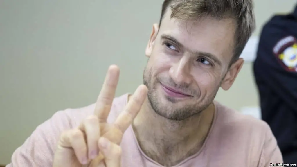A man with short light brown hair and stubble smiles while holding up his hand and making a peace sign with his fingers. He is wearing a light pink shirt. The background is blurred.