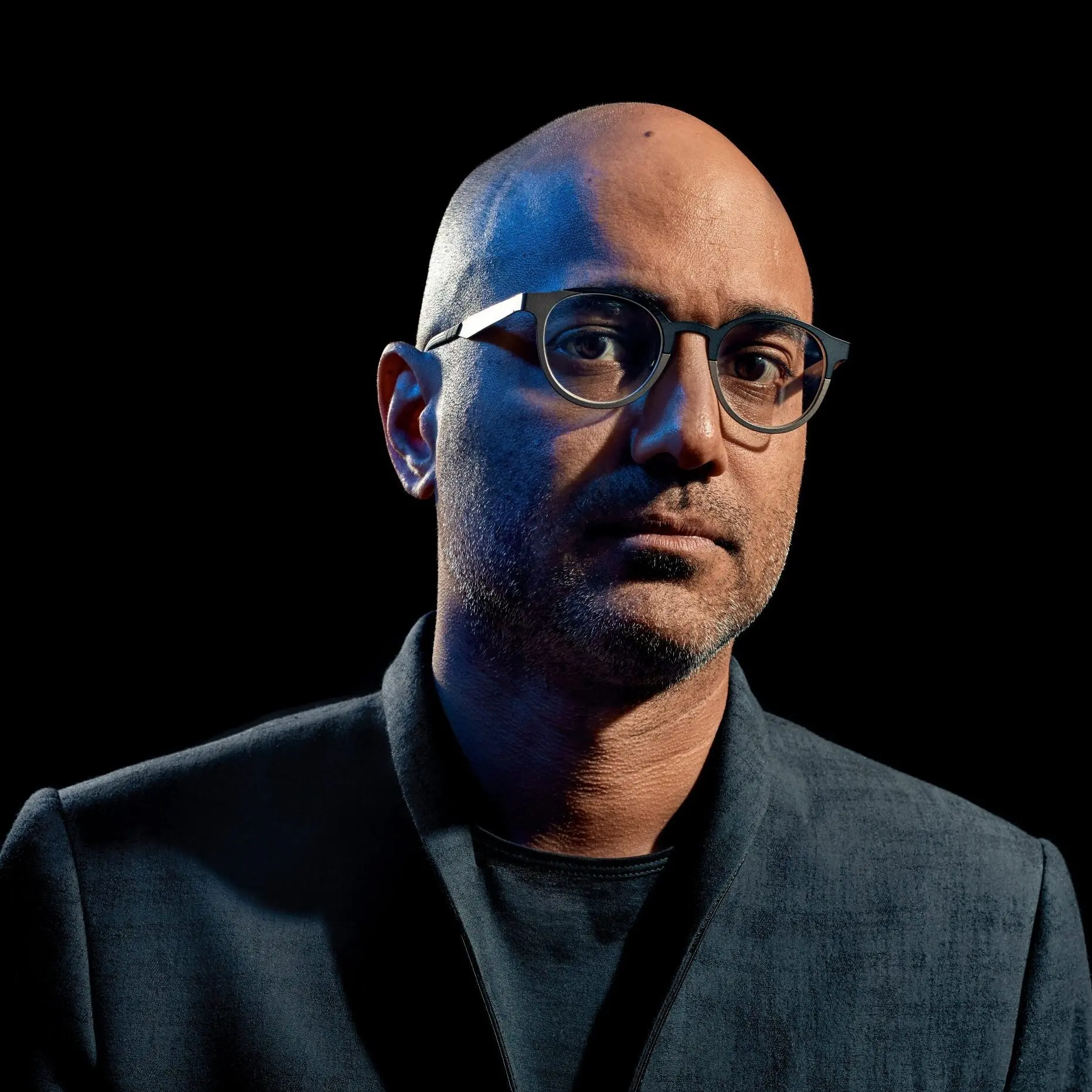 A bald man with glasses and a slight beard wears a dark jacket and shirt, looking directly at the camera against a black background, with blue light highlighting one side of his face.