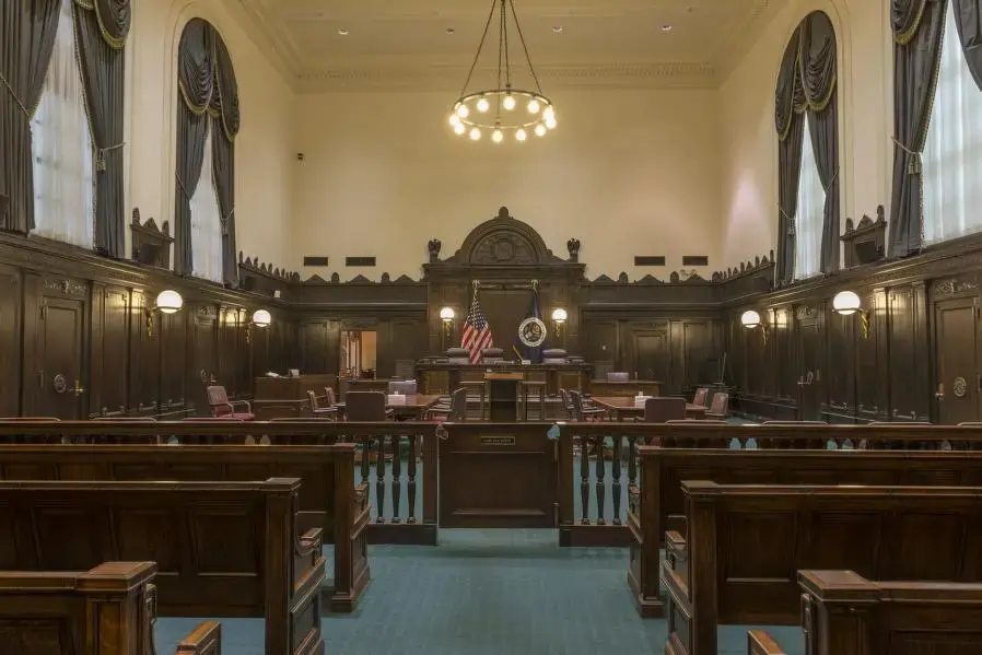 A stately, empty courtroom with dark wooden benches, a judges bench, witness stand, American flag, and large windows with tall curtains. A chandelier hangs from the high ceiling.