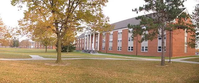 A large red-brick, two-story building with white columns stands on a grassy campus with trees and curving sidewalks, under an overcast sky.