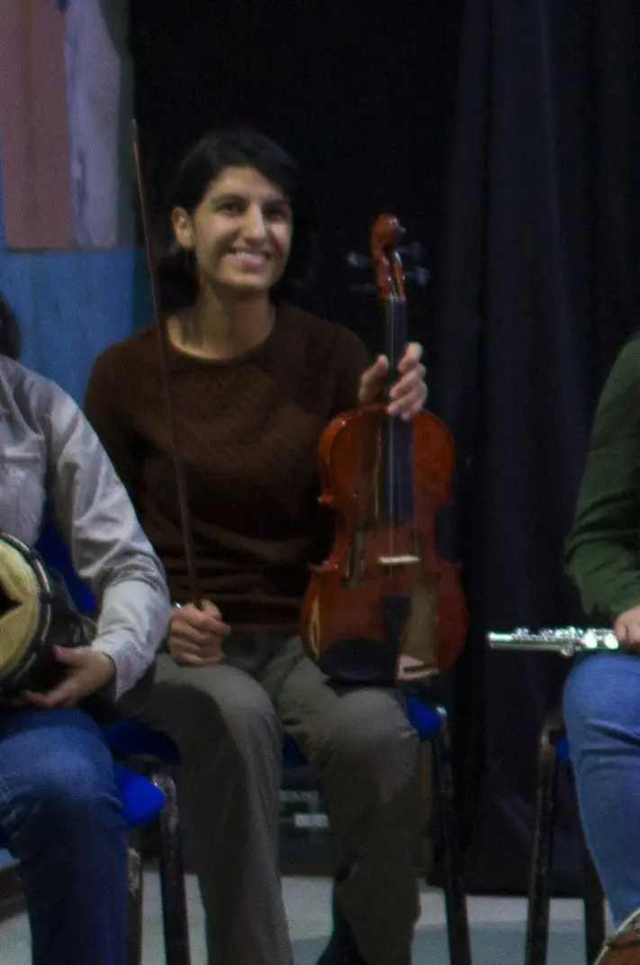 A woman sitting on a chair smiles while holding a violin in one hand and a bow in the other. She is indoors, with other people partially visible beside her.