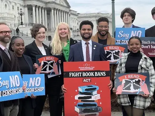 A diverse group of people stand outside a government building holding signs that say “Say NO to censorship” and “I read banned books,” surrounding a podium labeled “FIGHT BOOK BANS ACT.”.