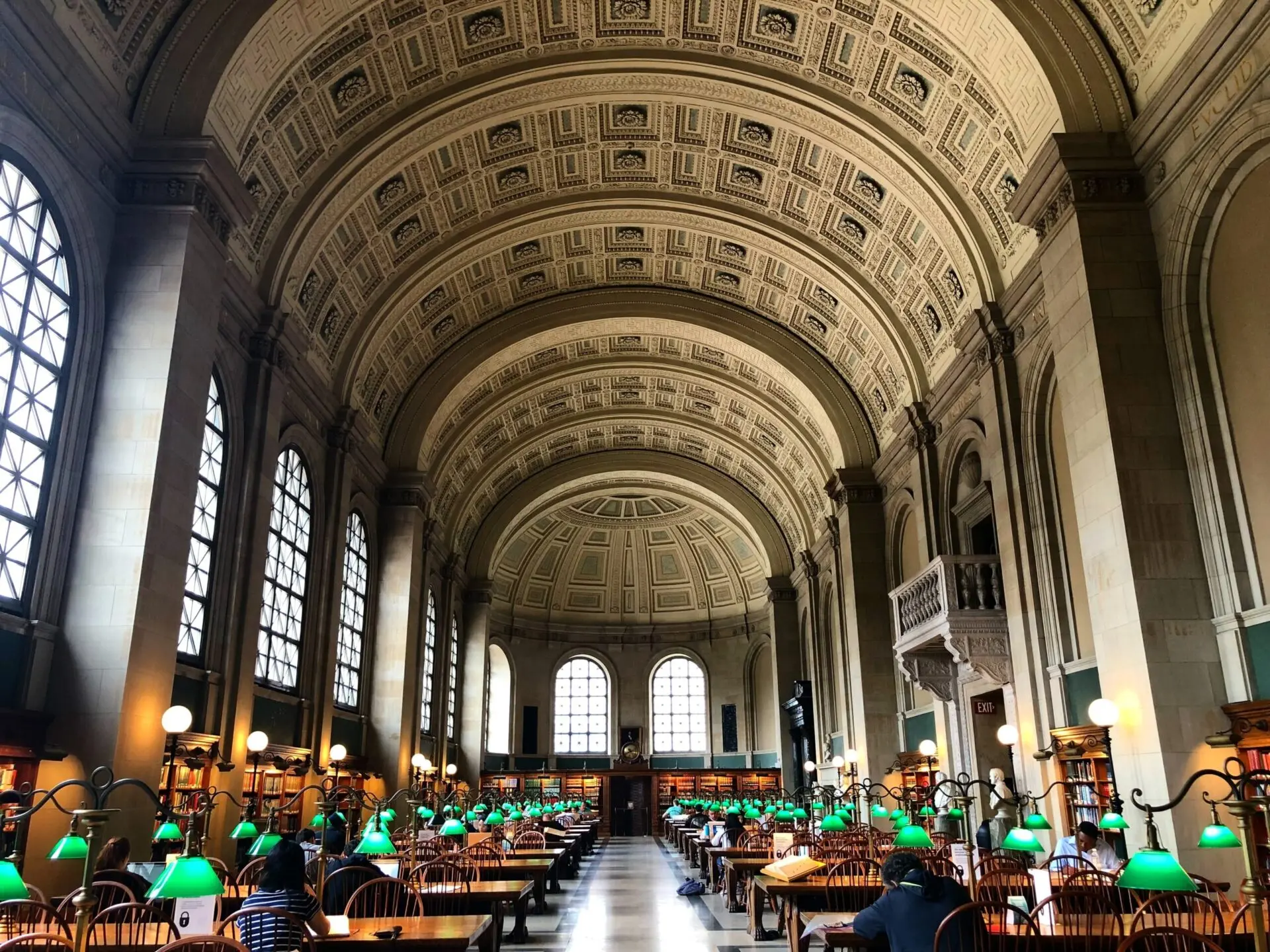 A grand library reading room with a high arched ceiling, large windows, and rows of wooden tables with green reading lamps. People are seated and reading in the well-lit, elegant space.
