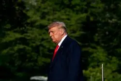 A man in a dark suit and red tie walks outdoors with a blurred green background of trees.