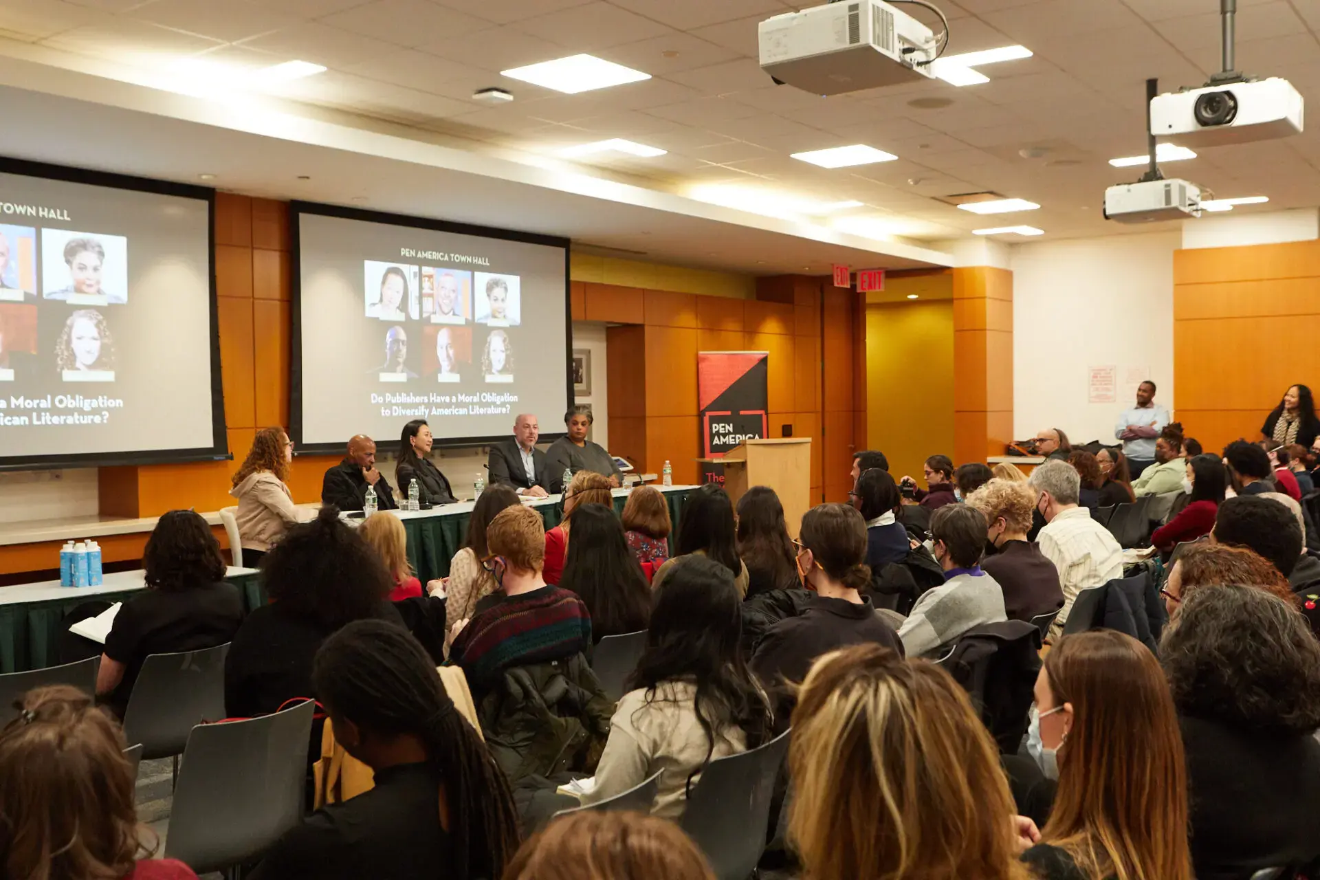 A diverse audience watches a panel of five speakers discussing on stage in a well-lit lecture hall. Two large screens behind them display the event title and speaker photos.