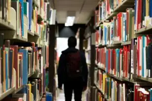 A person with a backpack stands between two tall bookshelves filled with books in a library, facing away from the camera toward the end of the aisle.
