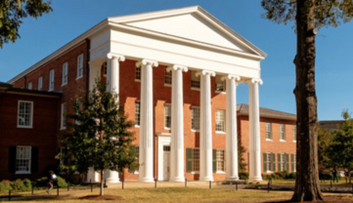 A large brick building with tall white columns and a triangular pediment, surrounded by trees and grass, under a clear blue sky.