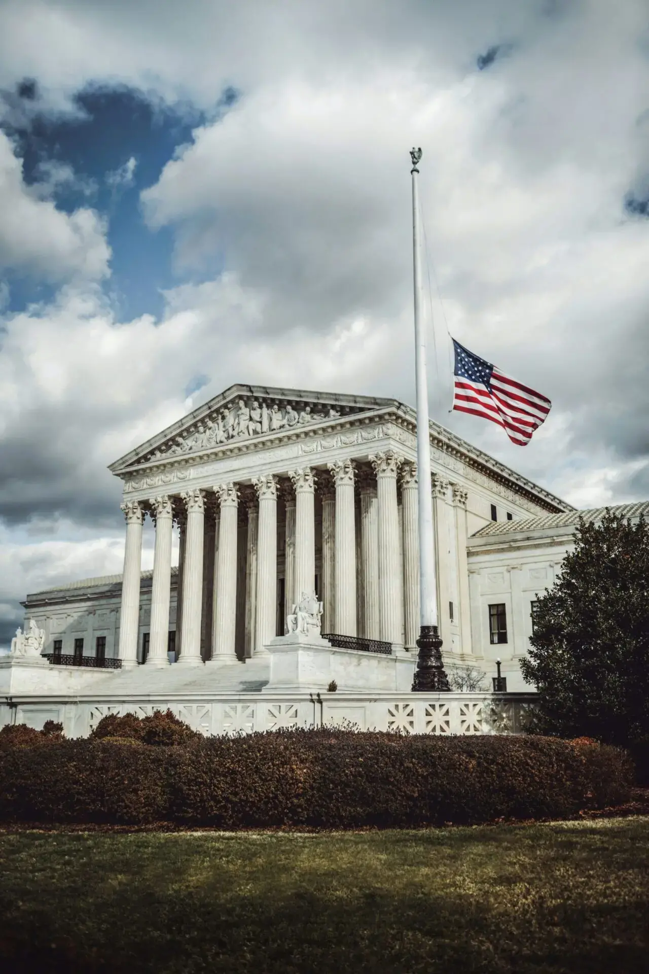 The American flag flies at half-mast in front of a large, white, classical building with columns, likely the United States Supreme Court, under a cloudy sky.