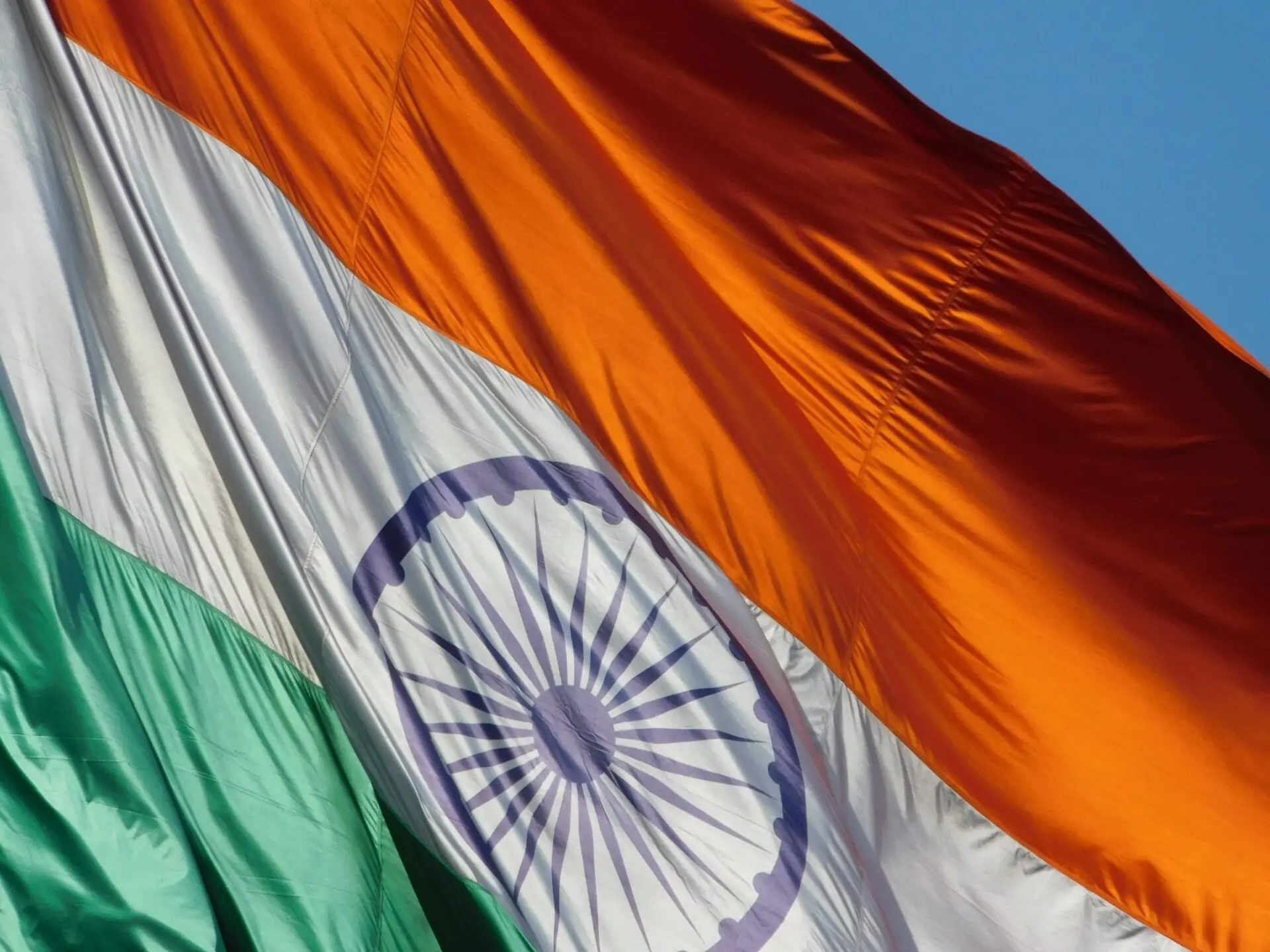Close-up of the Indian national flag, showing orange, white, and green horizontal stripes with a blue Ashoka Chakra in the center, waving against a clear blue sky.