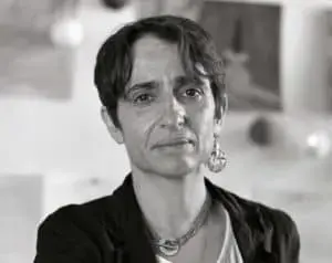 A person with short dark hair, wearing a dark jacket, light shirt, earrings, and a necklace, looks directly at the camera with a neutral expression. The background is blurred and appears to be an indoor setting.