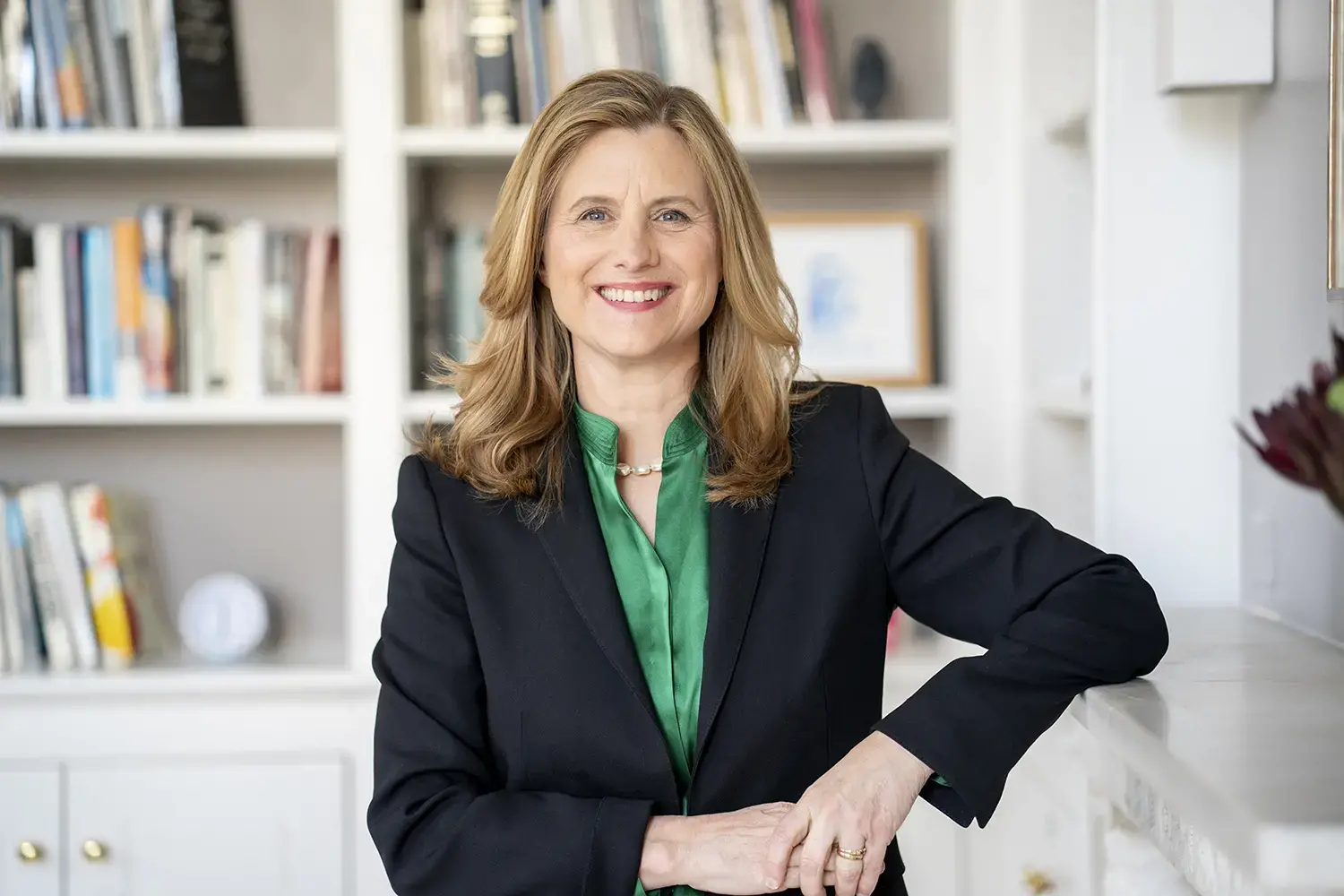 A smiling woman with blonde hair, wearing a black blazer and green blouse, stands indoors in front of white bookshelves filled with books and decor.