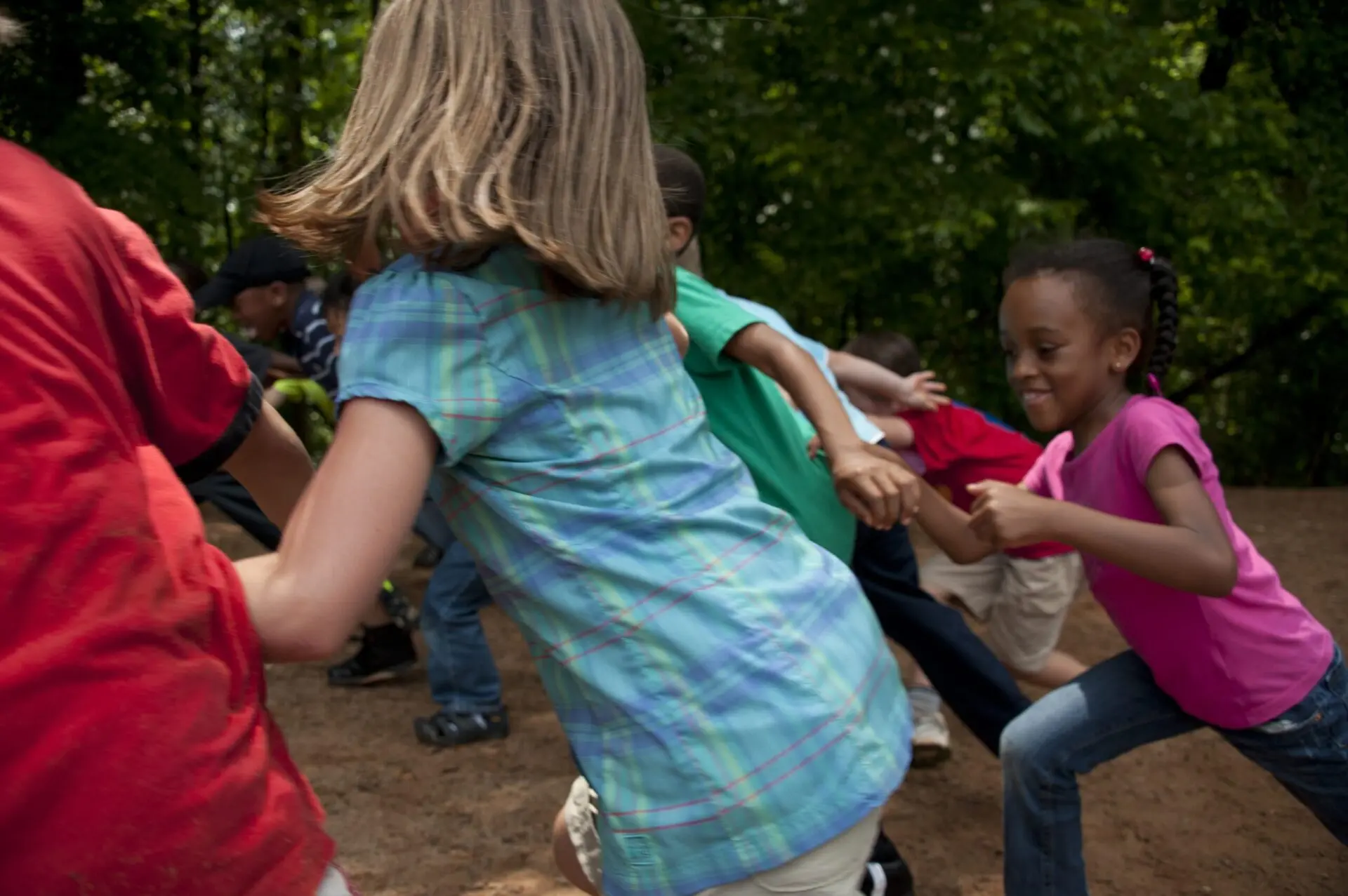 A group of children joyfully play together outside, holding hands as they run in a circle on a dirt surface, with trees and greenery in the background.