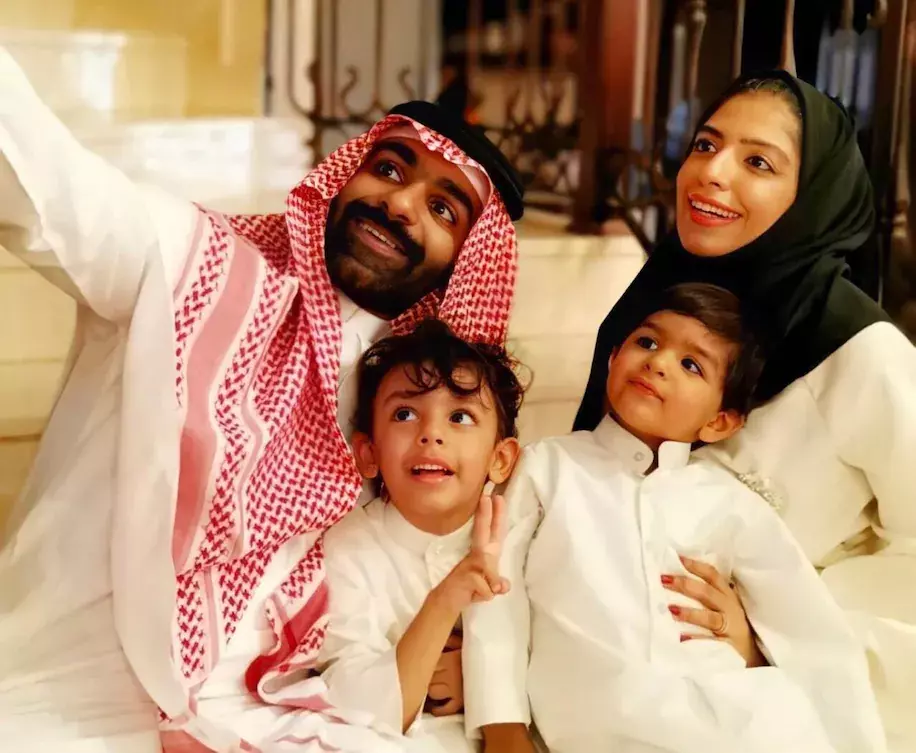 A smiling family of four, including a man in a red and white keffiyeh, a woman in a black hijab, and two young boys in white traditional clothing, pose together for a selfie indoors.