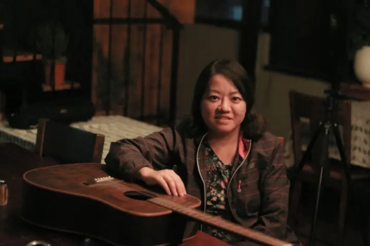A woman sits at a wooden table indoors, resting her arm on an acoustic guitar. She is wearing a dark jacket and smiling slightly at the camera. The background is dimly lit, with chairs and a potted plant visible.