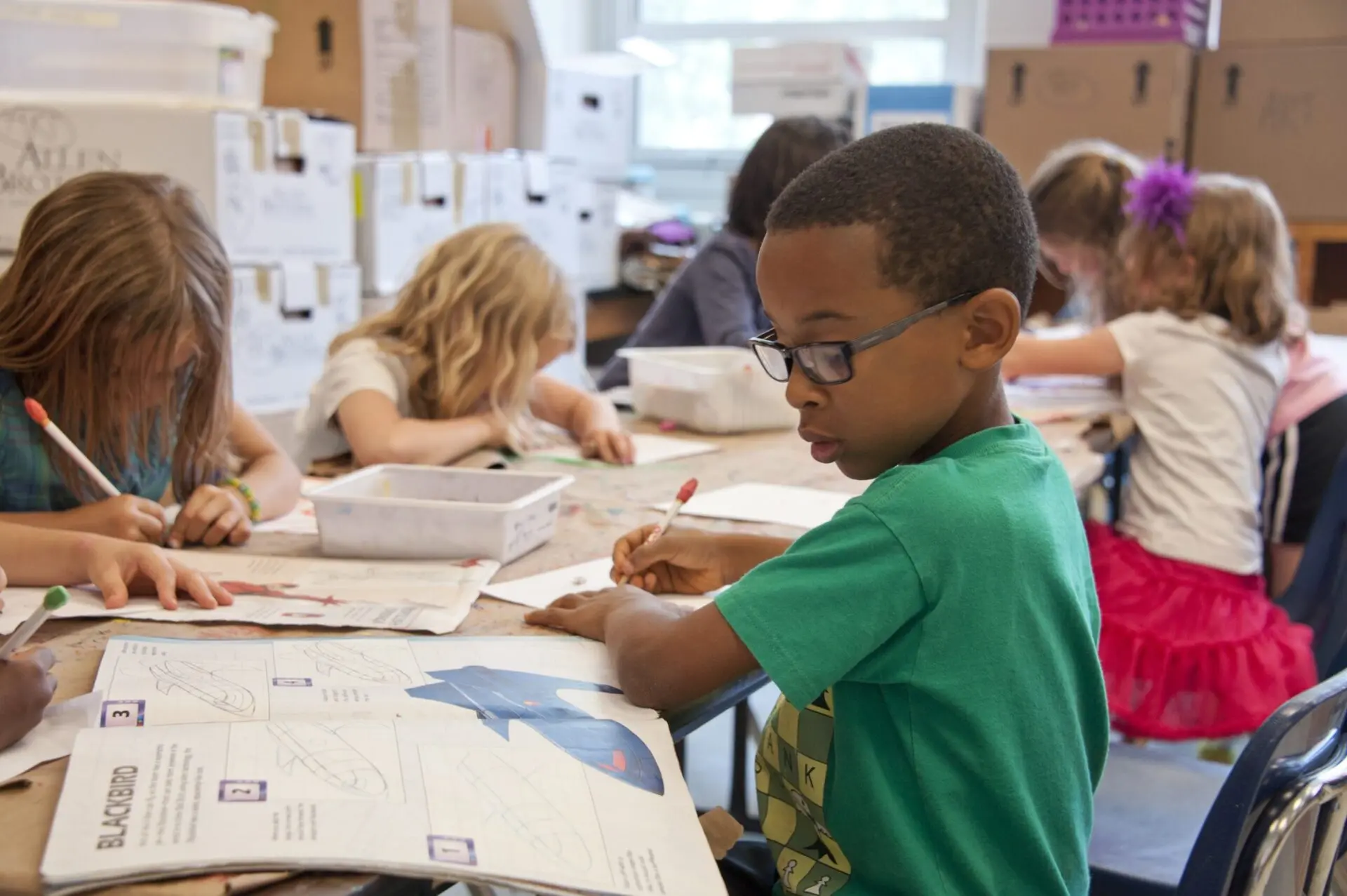 A group of young children sit at a table in a classroom, drawing and writing in workbooks. One boy in a green shirt and glasses focuses on his paper, while other children work beside him.