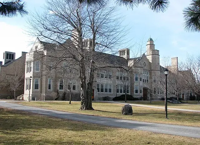 A large stone academic building with arched windows and a small tower, surrounded by leafless trees and a grassy lawn on a clear day. A paved walkway and lamppost are visible in front.