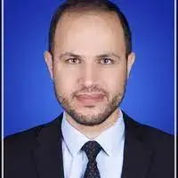 A man with short hair and a trimmed beard, wearing a dark suit, white shirt, and patterned tie, poses for a formal portrait against a solid blue background.