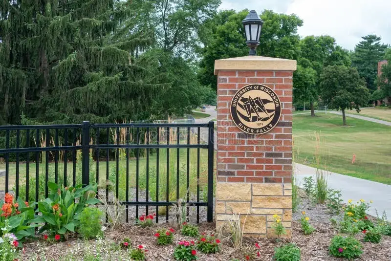 A brick pillar with a lamp and a University of Wisconsin-River Falls emblem stands next to a black fence, surrounded by colorful flowers and greenery on a campus walkway.