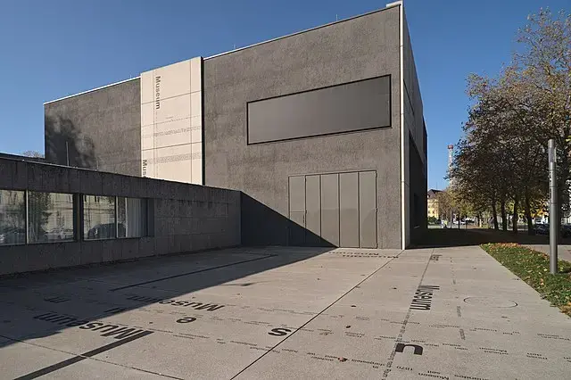 Modern, minimalist museum building with gray concrete walls, large rectangular and square panels, and a flat roof. The paved walkway in front has Museum written in various directions. Trees line the right side.