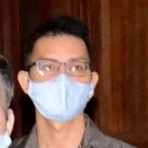 A person wearing eyeglasses and a light blue face mask stands indoors in front of a wooden background.