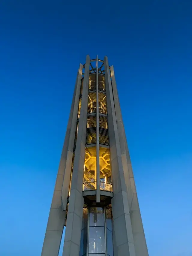 A tall, modern bell tower with illuminated interior lights stands against a clear blue evening sky, viewed from below.