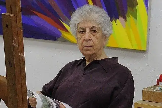 An older woman with short, curly gray hair sits holding a magazine in an art studio. She wears a dark shirt and looks seriously at the camera. Behind her is a colorful abstract painting.
