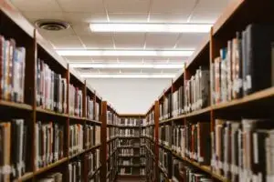 Rows of bookshelves filled with books line both sides of a library aisle, viewed from between the shelves. Fluorescent lights illuminate the ceiling, and the shelving continues into the distance.